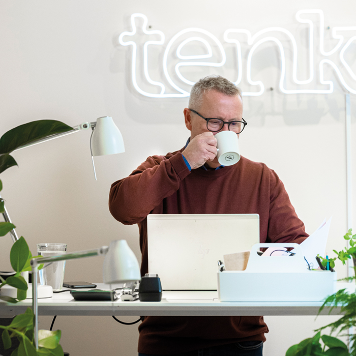 Svein Vekterli tar en slurk av en kaffekopp mens han står bak pulten og jobber på pcen. Foto